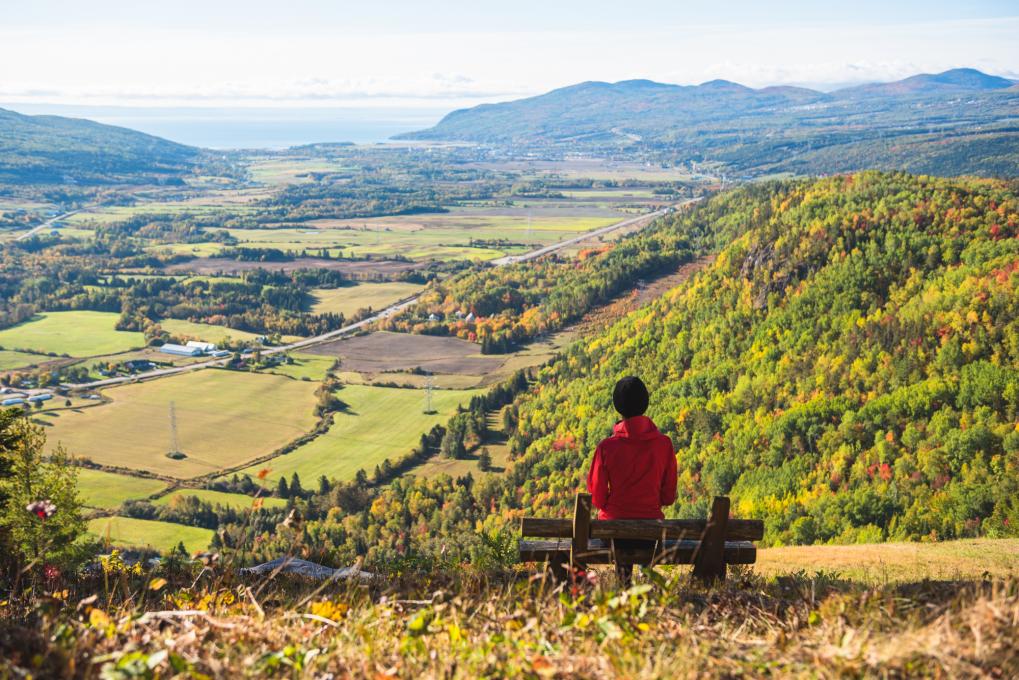 Une femme assise sur un banc au sommet d'une montagne observe le panorama et les couleurs d'automne dans la région de Charlevoix.